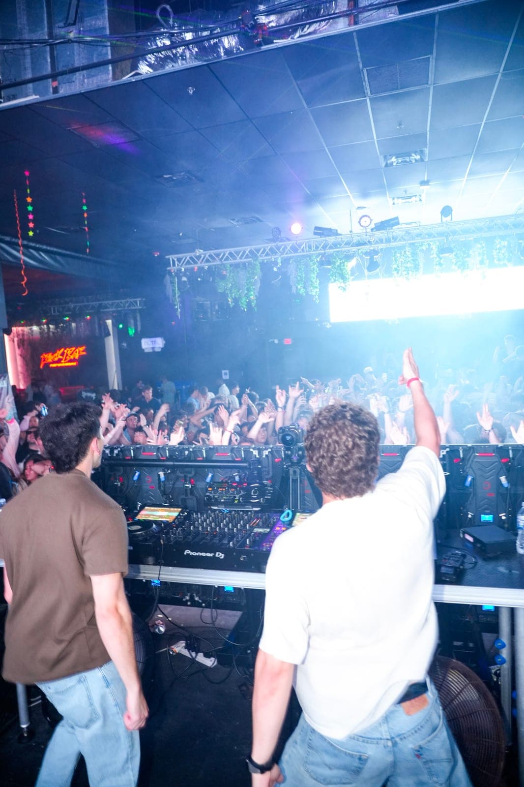 Two DJs at the booth, arm raised, facing a packed room under blue haze
